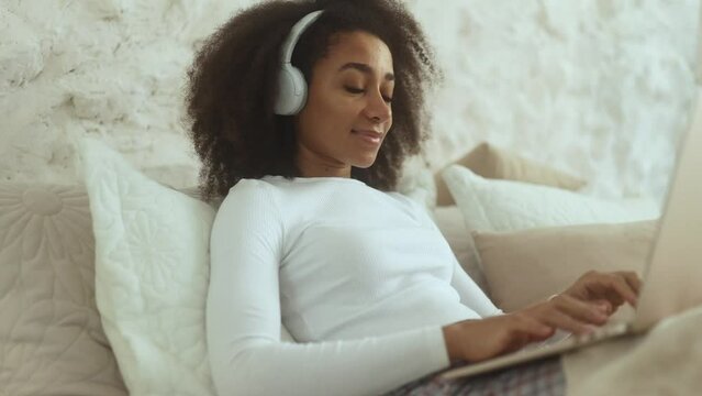 Charming curly woman relaxing at home listening music or working online browsing products in internet store reading news checking email or social media profile on laptop computer laying on bed indoors