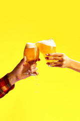 Male and female hand holds glass of cold light, foamy beer against vibrant yellow background.