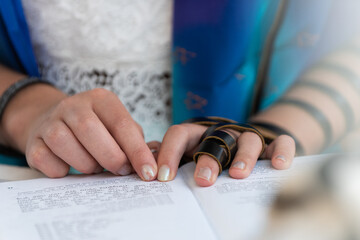 A young Jewish woman prays from a siddur or Jewish prayer book while wearing tefillin during her bat mitzvah ceremony. 