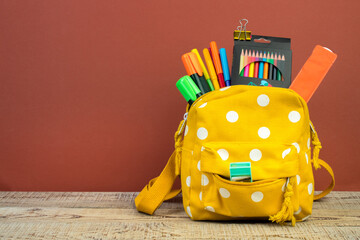 Yellow backpack with white polka dots with different colorful stationery on table. Brown...