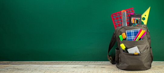 Backpack with different colorful stationery on table. Green background. Back to school. Banner...