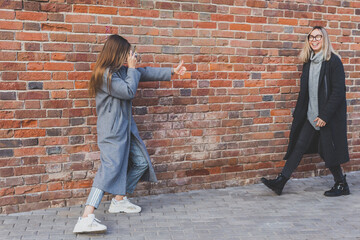 Fototapeta na wymiar Young millennial woman takes picture of her female friend in front of brick wall in city street - photographer and vintage camera hobby concept