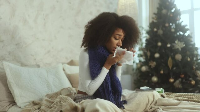 Sick curly woman sitting on bed with warm scarf coughing sneezing and having severe headache temperature during holidays at home Unhealthy female getting flu virus symptom near Christmas tree