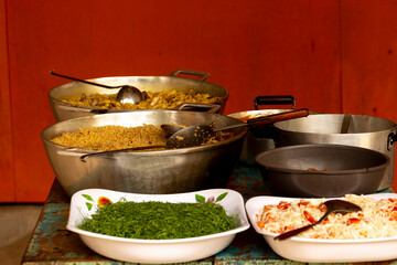 Mesa com diversos tipos de comida, panela de arroz, feijão, couve, salada de maionese e carne. 