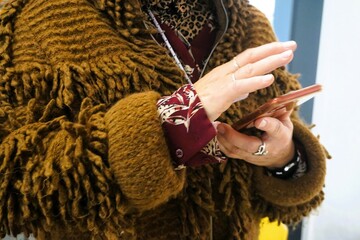 Oberkörper von Frau mit brauner Wollstrickjacke mit Fransen hält Handy in ihren Händen in Raum