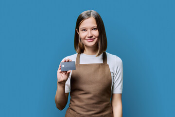 Young happy woman service worker in apron with credit card in hand, on blue background