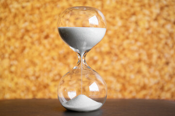 Hourglass with white sand dripping against gold color glitter background indoors.