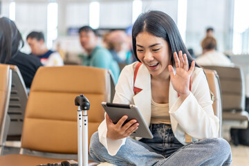 Attractive asian young woman traveler talking on video call to friend or family through tablet while waiting for her flight sitting on seat in departure gate at airport terminal.