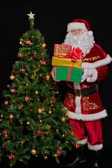 Santa Claus stands next to the Christmas tree and offers a boxes with a gifts. On the black background.