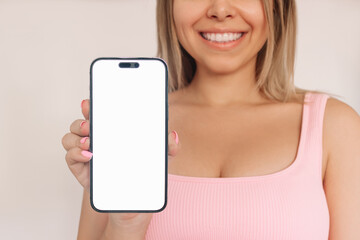 Cropped shot of a young smiling blonde woman holding mobile phone in her hand with white screen demonstrating copy empty space for text or design isolated on a light background. Mockup smartphone
