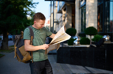Young traveller in a striped T-shirt walking on the street with a map and smartphone in hand and a...