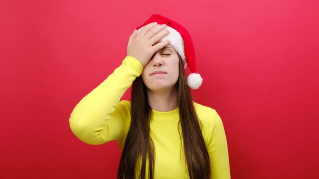 Sad young woman wearing yellow sweater and Santa Claus hat put hand on face facepalm epic fail omg gesture, posing isolated over pastel red studio background wall. Happy New Year 2024 holiday concept