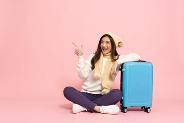 Happy Asian woman traveler sitting with luggage and pointing to copy space isolated on pink background, Tourist girl having cheerful holiday trip concept, Full body composition