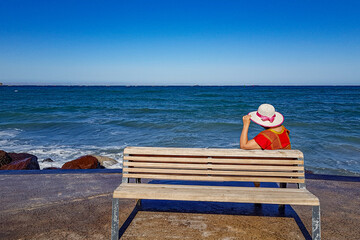 Rear view of an adult woman sitting on wooden bench on boardwalk or malecon with sea and horizon in background, hat and red blouse, sunny summer day in La Paz, Baja California Sur Mexico