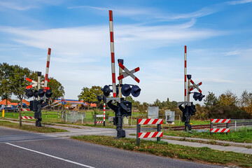 Railway crossing on a rural road, stop sign and warning lights, raised crossing barriers, farms and Dutch countryside in background, sunny autumn day in Weert, North Limburg, Netherlands