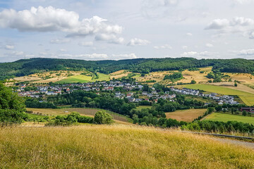 Fototapeta na wymiar Panoramic landscape of German countryside with small village and farmland, green leafy trees covering a hill in background, sunny day in Eiffel Kreis, Bitburg-Prüm district in Rijnland-Palts, Germany