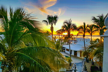 Coastal sunset seen from a terrace, palm trees against reddish blue and orange sky, sun beginning to set in background, calm day in La Paz, Baja California Sur Mexico