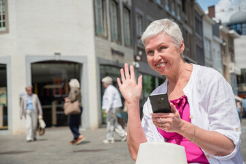 a sixty year old caucasian woman in a hat and stylish summer clothes waves to a passerby and smiles and holds a smartphone in his other hand, sits on a bench in the city, High quality photo - obrazy, fototapety, plakaty
