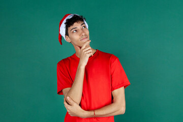 Chico pensativo vestido de rojo y con gorro de papa noel. Hombre en navidad. Fondo verde