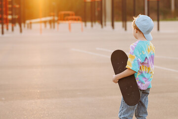 Child with a black skateboard in an empty skate park at sunset. In a bright putt shirt, jeans and a cap, he holds a skateboard with one hand. Active recreation or skate school concept with copy space.