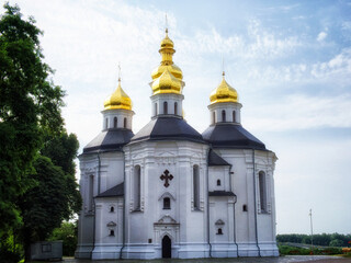 Fototapeta na wymiar A white church with golden domes and crosses, nestled amidst lush greenery under a blue sky with clouds. Beautiful sky over the Orthodox Church of St. Catherine in the Ukrainian city of Chernihiv.