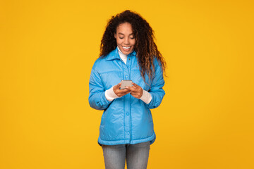 Young black woman in blue jacket using smartphone on yellow background