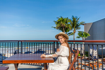 Fototapeta na wymiar Seaside elegance. Woman in hat at wooden table taking in breathtaking beach vista. Swirling ocean waves meet palm-adorned skyline.