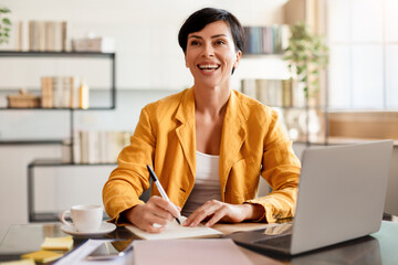 Smiling Middle Aged Businesswoman At Laptop Taking Notes In Office - obrazy, fototapety, plakaty