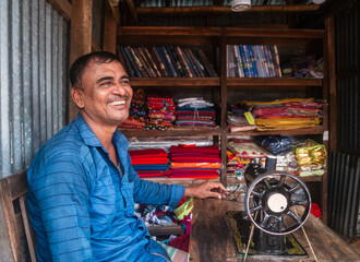 Smiling portrait of a south asian rural tailor sitting in front of his old fashioned sewing machine inside his small clothing store 