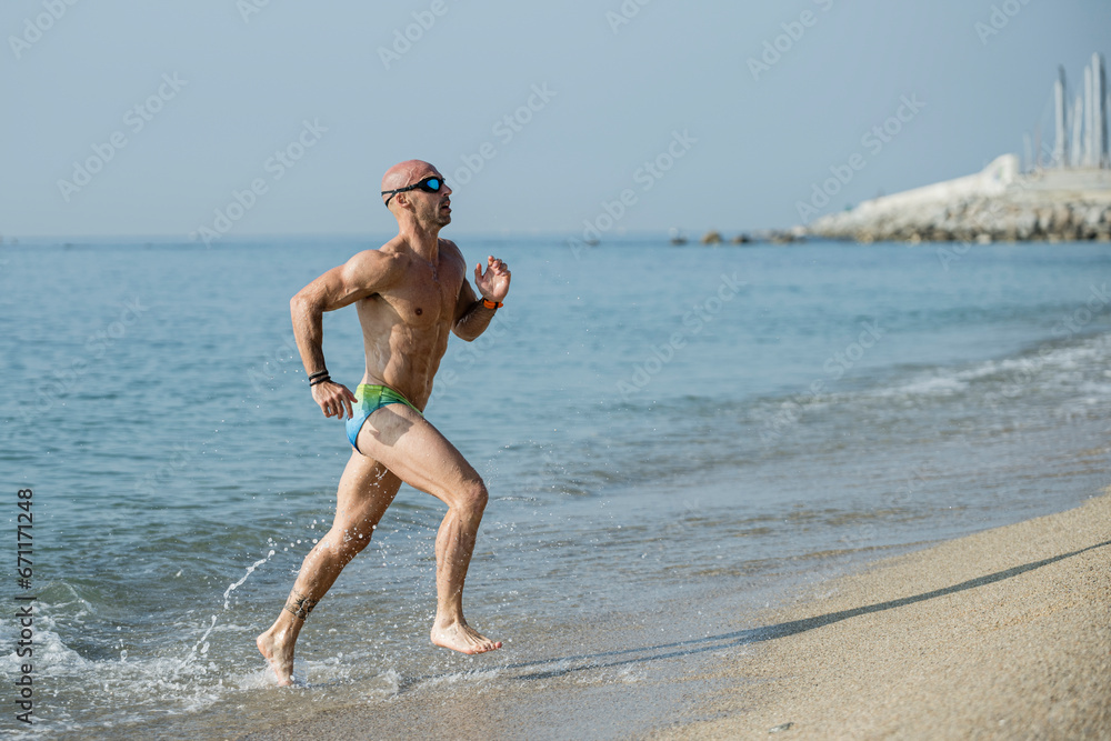 Wall mural side view of a sexy muscular man in colorful speedo running out of the sea. professional male athlet - Wall murals