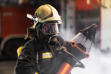 Fireman portrait wearing full equipment and oxygen mask. Firefighter with power hydraulic cutting tool. Smoke and fire trucks in the background.