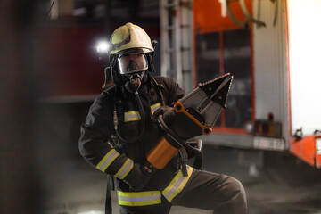 Fototapeta premium Fireman portrait wearing full equipment and oxygen mask. Firefighter with power hydraulic cutting tool. Smoke and fire trucks in the background.