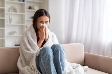 Sick Asian Woman Blowing Nose In Paper Tissue While Sitting On Couch