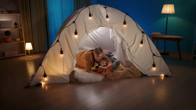 A father and son are reading a book in a tent set up in the living room. A man and a boy lie on their bellies on a soft fur plaid in an embrace, the son is listening to his father, holding a soft toy.