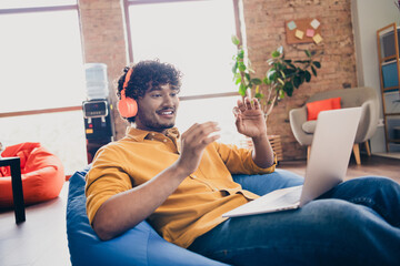 Photo portrait of attractive young man wear yellow shirt sit blue bean bag talk video call headphones modern workplace room home design