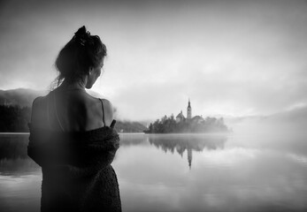 Woman in gray coat watching a sunrise among the fog on the shores of Lake Bled, Slovenia II - obrazy, fototapety, plakaty