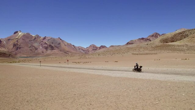 side view man on motorcycle followed by drone discovering a lonely desert road with mountains and lagoons 