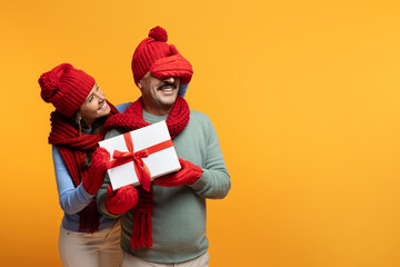 Smiling senior european wife in hat closes eyes with hand to husband at winter, gift box present