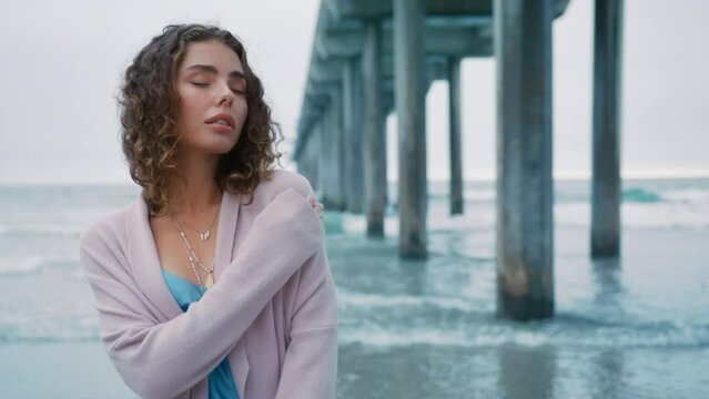 Sexy young girl on gloomy coastline. Stylish attractive woman enjoy summer vacation 4K RED. Luxurious curly lady walking in front of ocean pier on beach wearing blue slip silk dress and beige cardigan