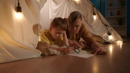 Dad and son drawing together with colored pencils in a notebook, a man is helping the child. Dad and son are lying on their stomachs in a garland tent that is in the living room close up.