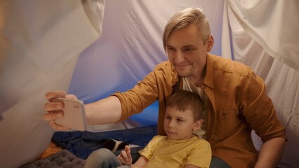 Obraz premium A father and son sit on fur skins in a tent in a dark living room and use a smartphone. Man and boy take selfies together, smiling, close up.