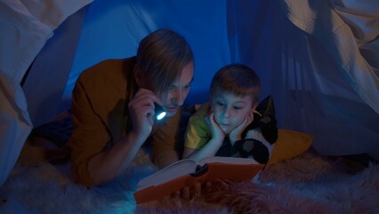 Obraz na płótnie Canvas Dad and son reading a book in a tent in a dark room close up. A man and a boy lie on their bellies on a soft fur plaid, the man holds a flashlight pointing it at the book, the son nearby listening.