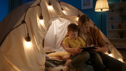 Father and son use a tablet, play an online game together, watch a photo, video, cartoon. Son sits on father's lap, near garland tent set up in dark living room close up.