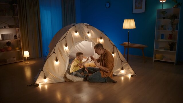 A dad is teaching his son to count. Man shows fingers on hands, that boy is counting. Father sits on floor near tent in living room where boy is sitting. Concept of parenting, teaching.