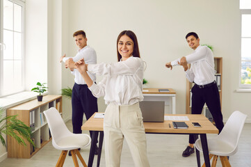 Happy corporate business team having fitness workout in office. Group of three people doing sports exercises. Smiling young woman and two men standing in office and doing shoulder stretching movements