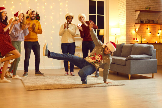Happy young multiethnic people having fun at Christmas party in beautiful modern studio apartment. Group of excited millennial friends in Santa hats applauding funny dancing guy in ugly Xmas sweater