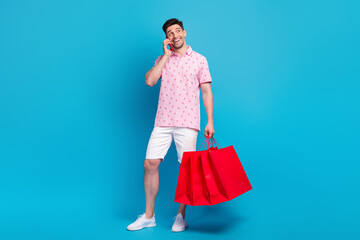 Full length photo of cheerful nice guy hold mall bags speak communicate telephone isolated on blue color background