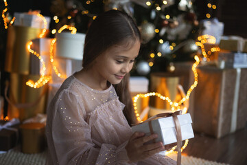 Cute little girl with long hair in dress sitting on the floor and opens a box with a present for background Christmas tree. Xmas holiday concept