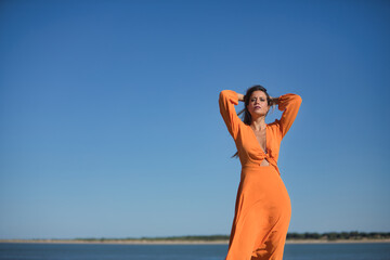 Young and beautiful woman in an orange dress, with her hands on her head, in sensual attitude, on the beach, with the wind blowing through her hair and dress. Concept beauty, fashion, trend, air.