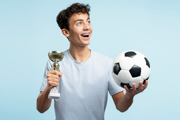 Portrait of positive teenager football player with dental braces holding trophy cup and soccer ball...